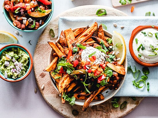 Delicious sweet potato fries topped with guacamole, peppers, and fresh herbs on a wooden board.