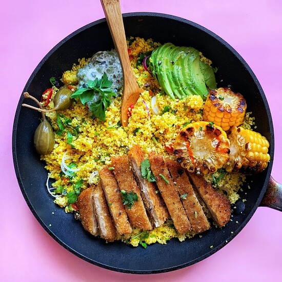 Delicious couscous dish with avocado, grilled corn, crispy protein, and vibrant garnishes in a black pan.